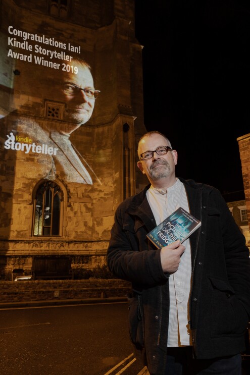 Ian W. Sainsbury holding his book in front of a Storyteller projection of his head shot congratulating him on his award.