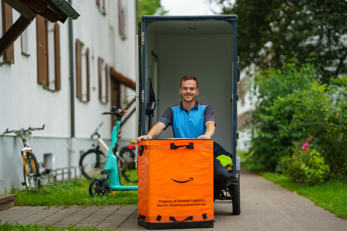 Enis sizt lächelnd auf der Ladefläche des E-Cargo-Bikes. Vor ihm steht eine große orange-farbene Amazon-Versandtasche. Links sieht man die Umrisse eines Mehrparteienhauses und davor Fahrräder und einen E-Roller.