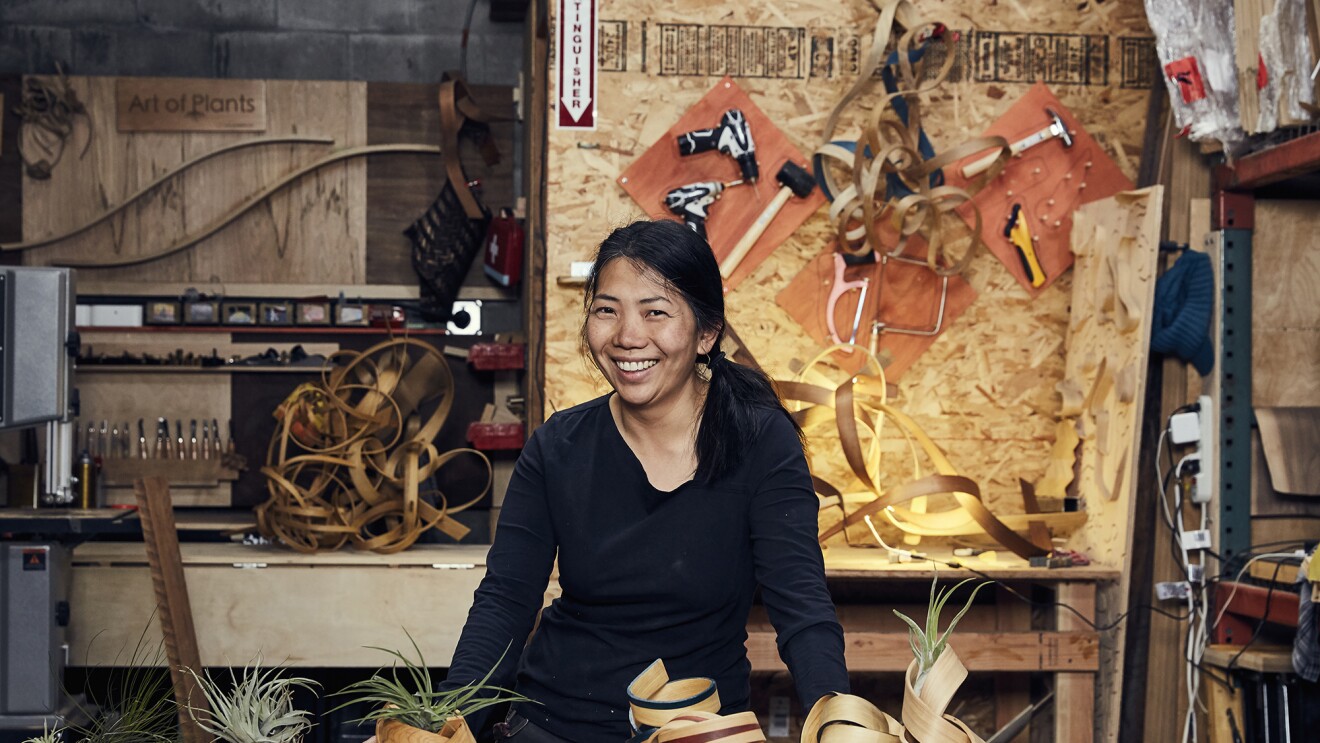 一个女人站在工作台后，对着镜头微笑。在她面前是几个手工制作的弯曲木材空气植物容器，在她身后坐着更多的木材和工具。