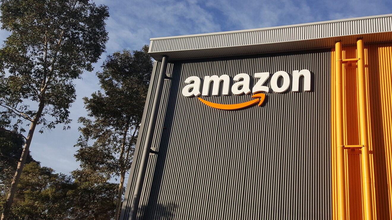 An image of the outside of an Amazon fulfilment centre. The Amazon logo is on the side of the gray building and there are trees and a blue sky in the background.