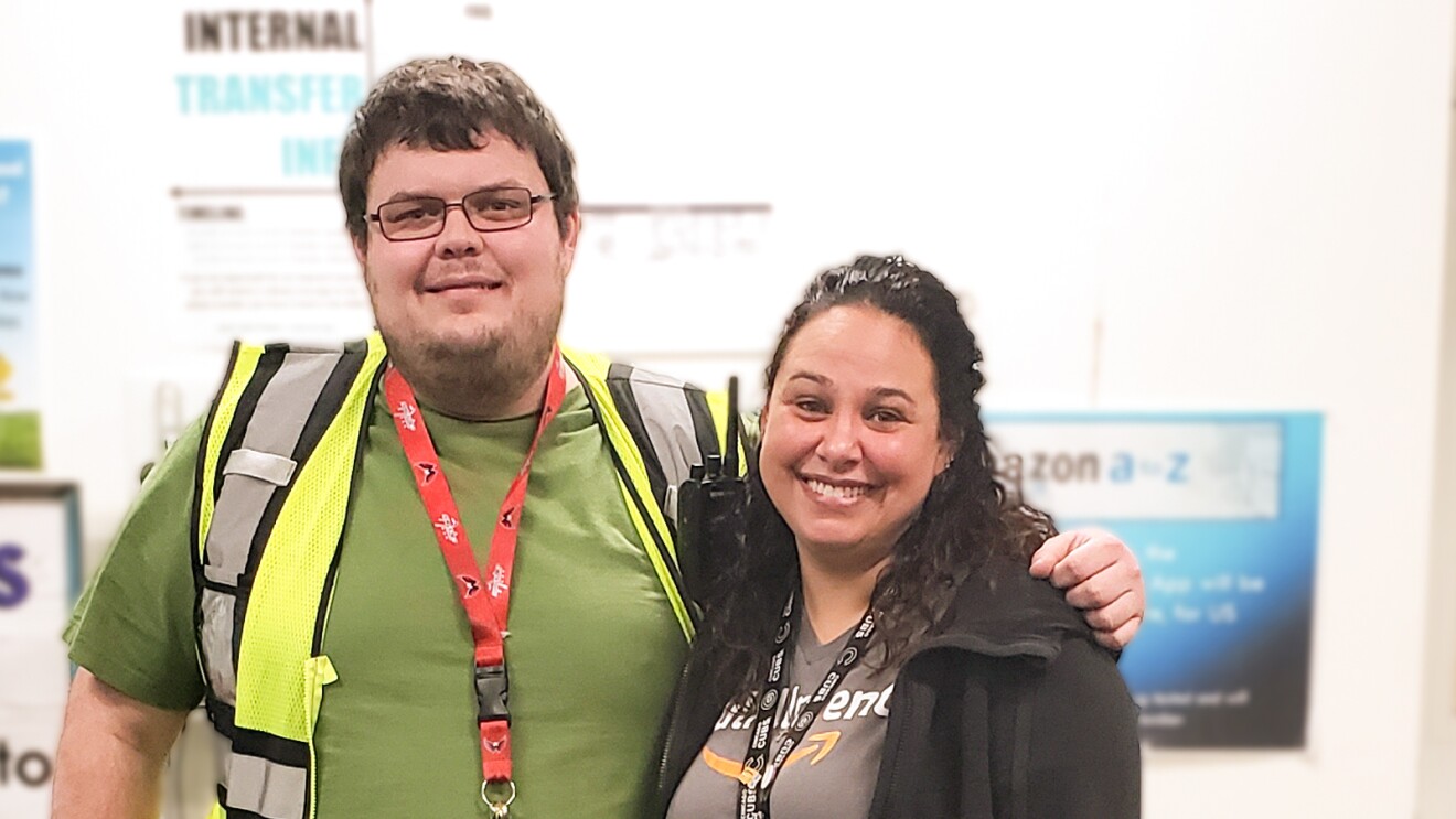 A woman and a man pose together for a photograph. The man wears a green T-shirt and a yellow safety vest. The woman wears a black fleece jacket and a gray T-shirt with the Amazon smile logo.