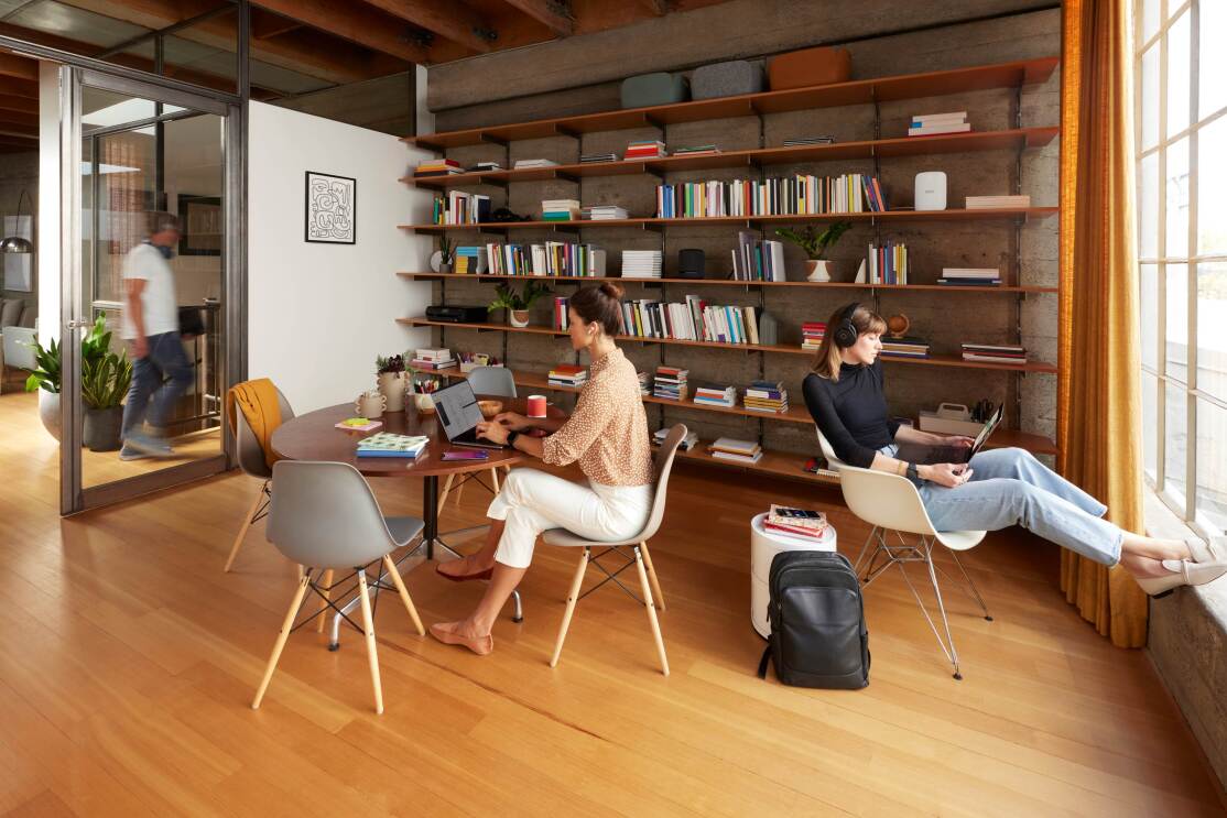 多数の本が並べられているオフィスのような空間で、女性1人がヘッドセットをかけ、パソコンを操作しながら窓を向け座っている。もう1人の女性はテーブルでパソコンを操作している。