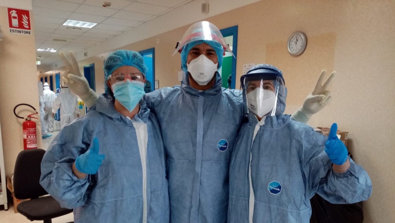 Tre persone in un ospedale indossano tute protettive e dispositivi di protezione personale come mascherine e guanti. Guardano verso la fotocamera e salutano.