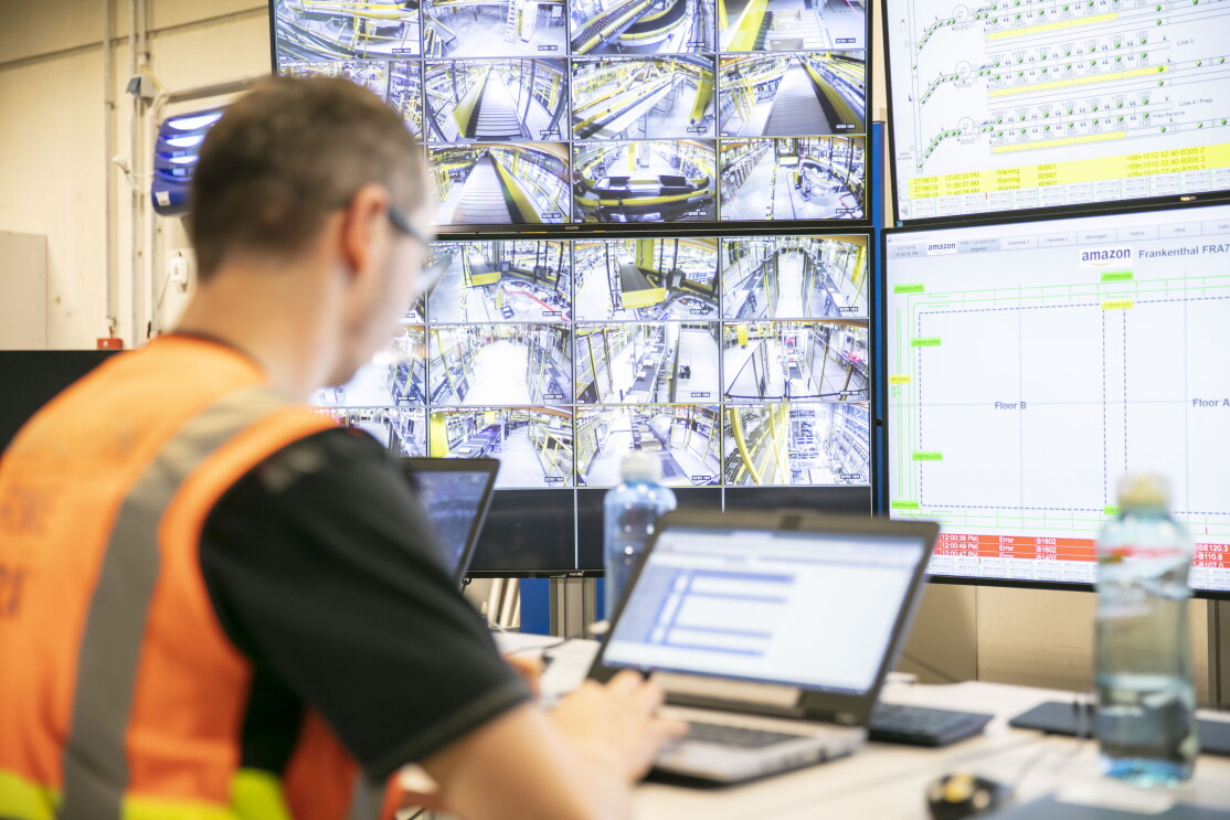 Ein Mitarbeiter in orange-farbenen Weste tippt an einem Laptop. Vor ihm sieht man auf großen Bildschirmen Technik und Förderbänder.