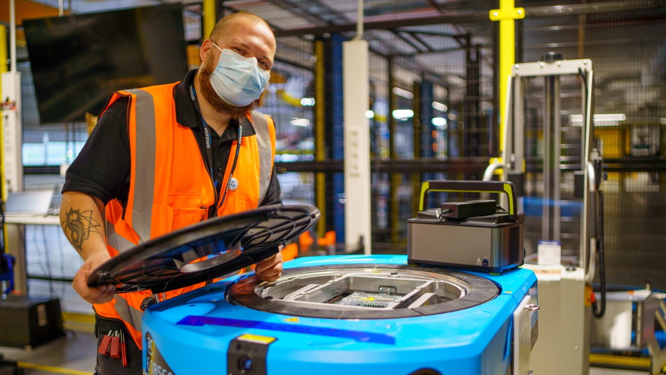 Logistik Mitarbeiter arbeitet an einem Roboter, er trägt eine orangene Warnweste und einen Corona-Mundschutz