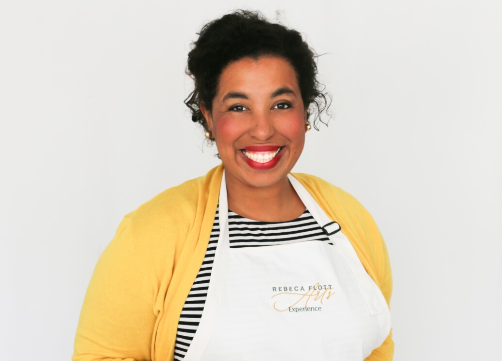 An image of a woman smiling for a photo against a white background. She is wearing a yellow shirt and a white apron.