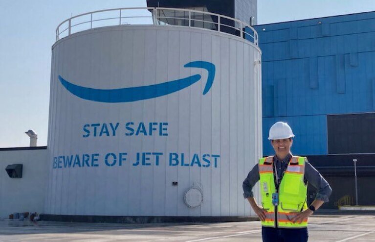 An image of Sarah Rhoads smiling in front of a large, white structure that says "Stay safe, beware of jet blast" at an Amazon Air Hub. She is wearing a safety vest and a hard hat.