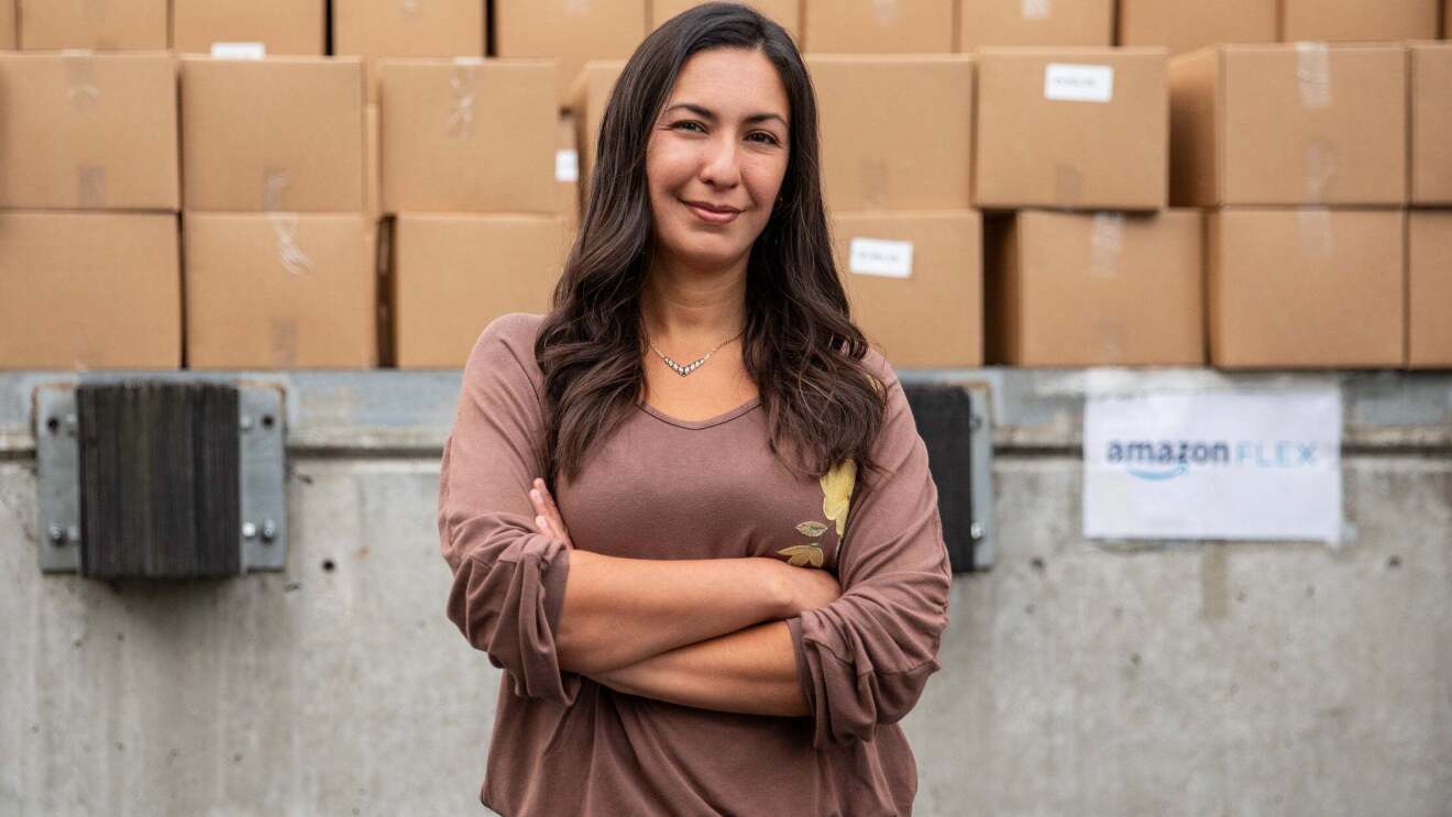 A woman stands front of a cement wall, stacked with boxes, and with a sign that says "Amazon FLEX" She crosses her eyes and smiles at the camera.
