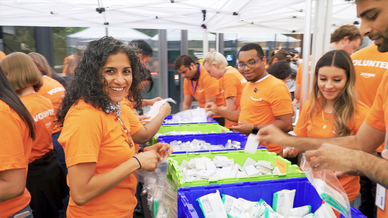 A group of Amazon employee volunteers wear matching orange T-shirts and pack Ziploc bags with supplies.