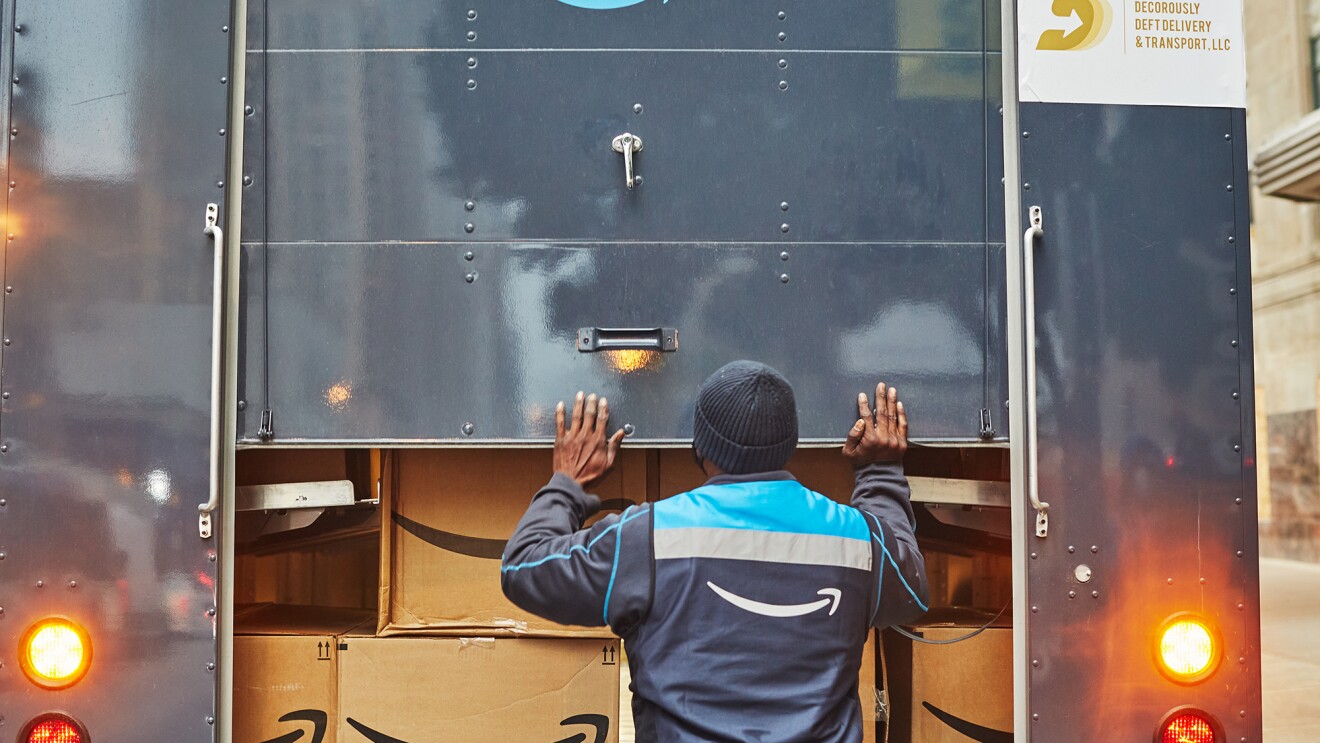 An image of a man closing the back of an Amazon delivery truck that is loaded with Amazon boxes inside of it.