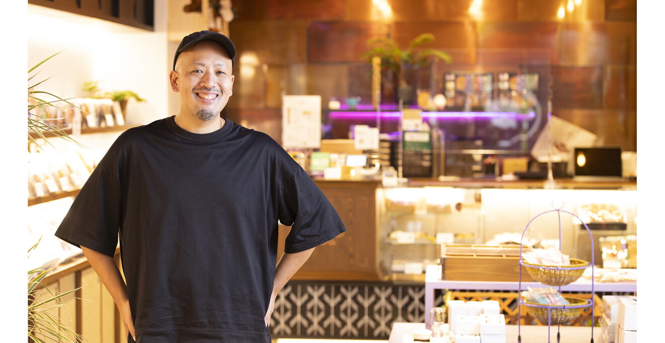 店舗の前でTシャツとキャップのスタイルで笑顔の松﨑さん