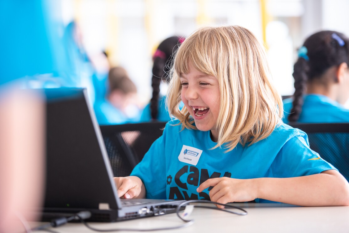 Ein blondes Mädchen im blauen Camp-Amazon-Shirt sitzt vor einem Laptop und lacht