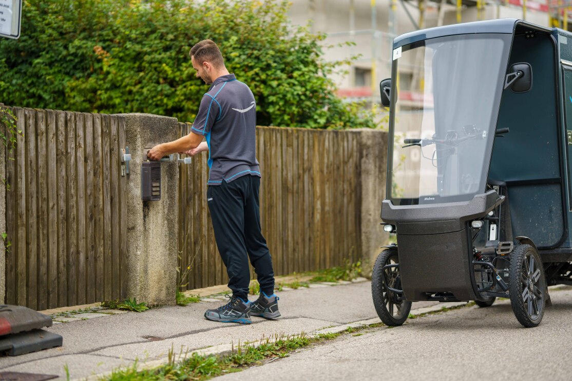 Enis von hinten vor einem Briefkasten, der von einem Holzzaun umgeben ist: Man sieht ihn von hinten, auf dem Shirt ist ein großes Amazon-Logo zu erkennen. Rechts neben ihm steht sein E-Lastenrad.