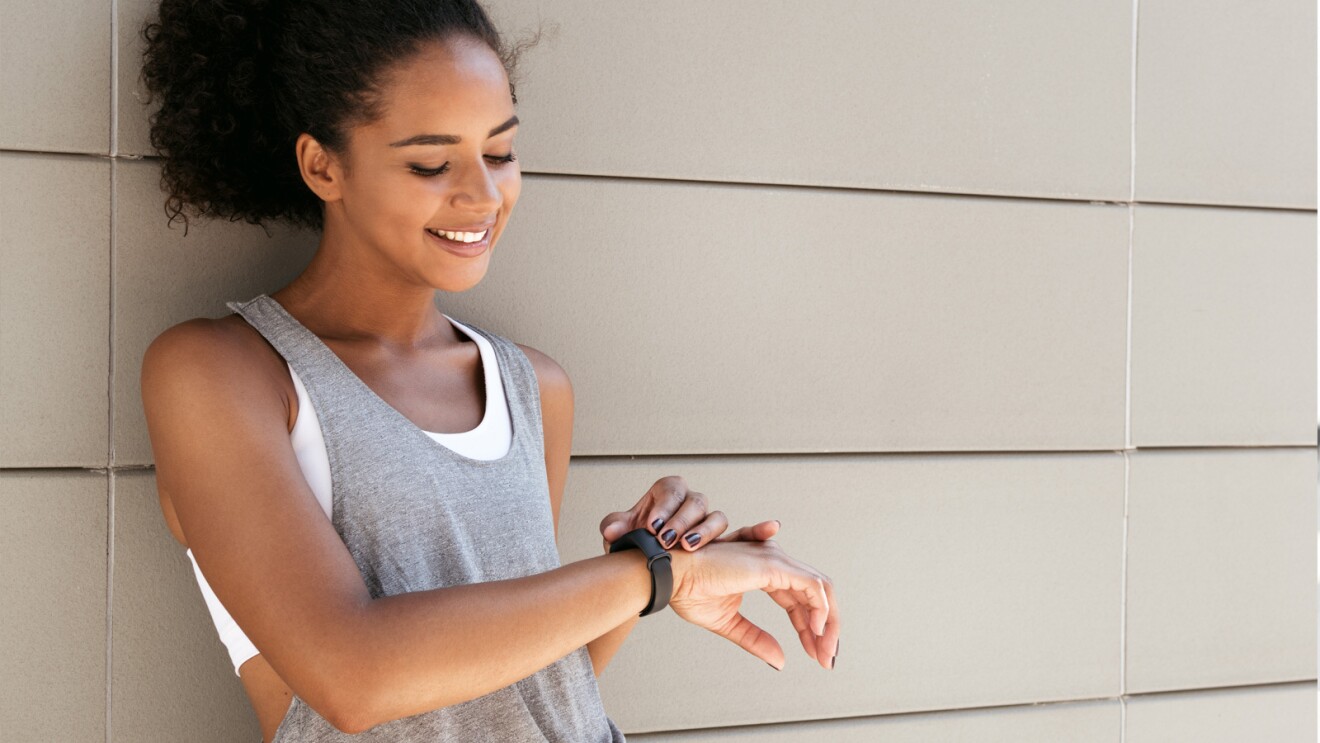 A woman leans against a wall checking her fitness tracker on her wrist. 