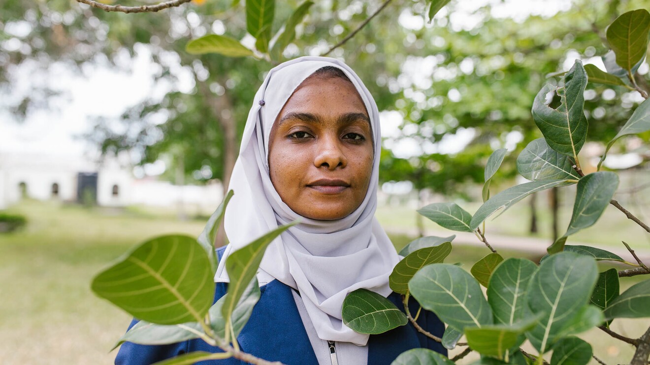 A young woman looks at the camera, from behind leaves on a tree branch