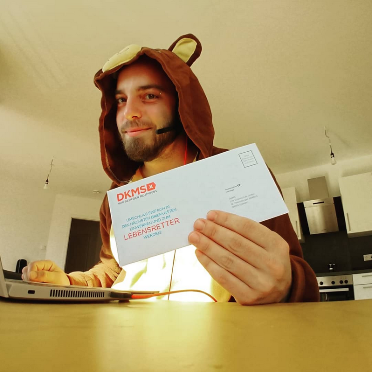 Ein Mann mit Teddybärenkostüm sitzt vor einem Laptop und hält einen Zettel der DKMS in die Kamera. Er lächelt.