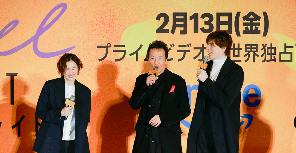 オレンジ色の壁の前、オレンジのステージの上に、黒い服を着て横一列に並んだ3人の人物。全員マイクを手に持って会話をしている。