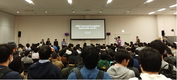 広いホールに集まった参加者たちの後ろ姿。奥の壁に大きなモニター。参加者たちは並んで座り、モニターの方を向いている。