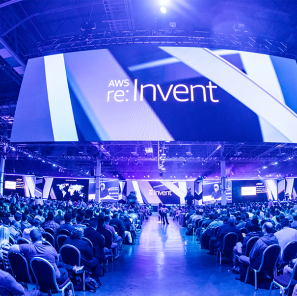 AWS re:Invent conference, view shows hundreds of individuals sitting in chairs at a conference. A huge screen hangs above them, the space is awash in blue light.