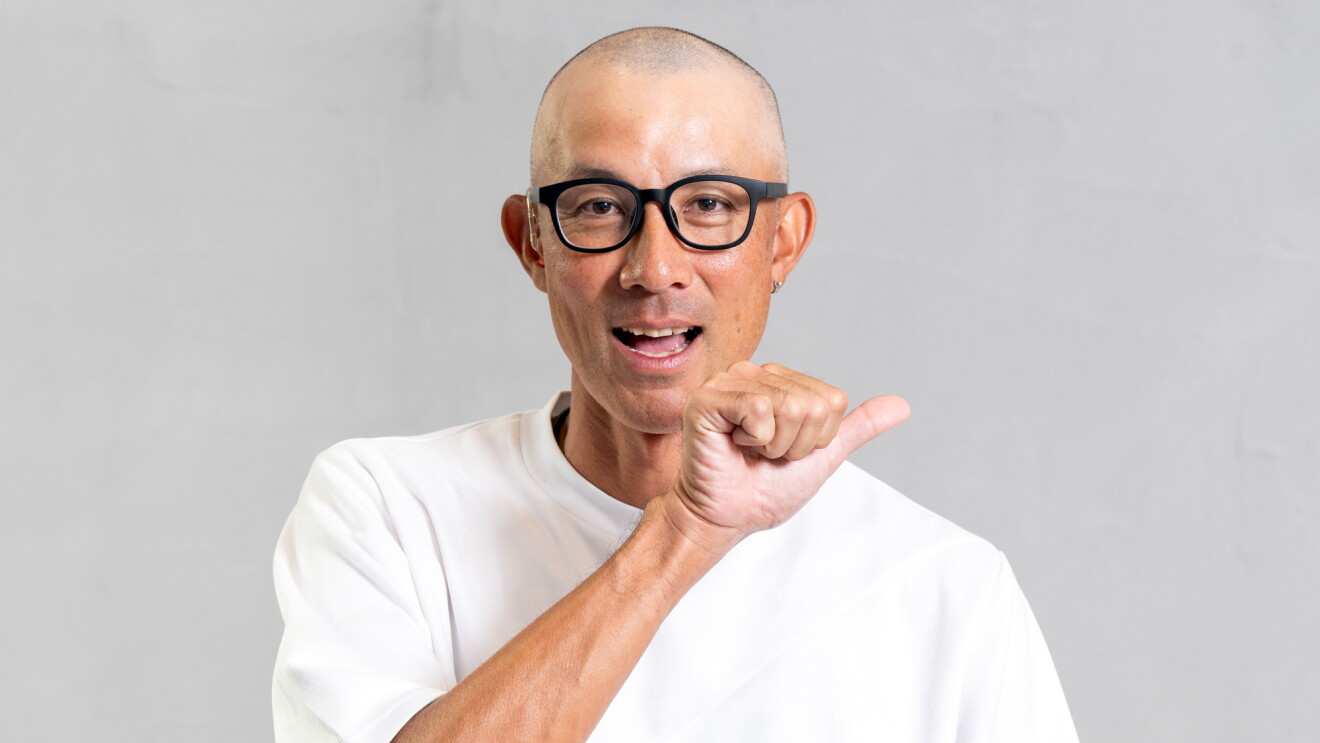 白いTシャツを着た男性が顔の前に親指だけを広げほかの指は握った形を作っている。口元は開いている