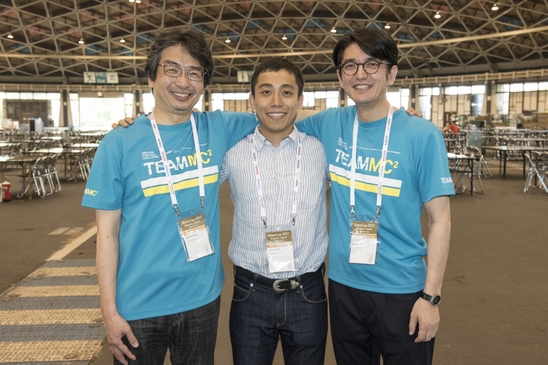 屋外ホールのような広い場所で肩を組んで並ぶ3人の男性。中央は青と白のストライプシャツ、左右の2人は水色の揃いのTシャツを着用。全員首から参加証のようなカードを下げている。