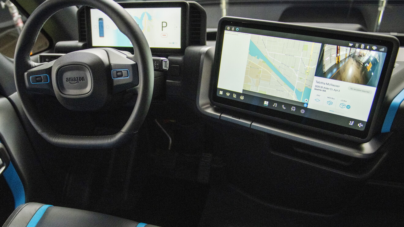 Primer plano del volante y de la pantalla de navegación. A la derecha el volante de color negro con el logo de Amazon y una pantalla con la información técnica relacionada con el coche. A la derecha una pantalla de navegación con un mapa y una fotografía del destino. 