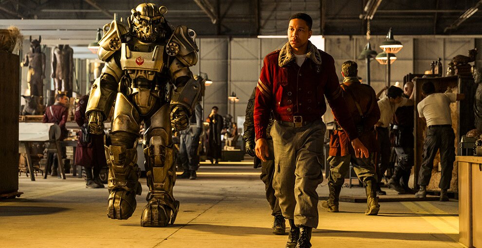 工場のように武骨で殺風景な屋内を歩く赤いジャケットの人物。左側には全身銀色のパワーアーマーもしくはロボットが並んで歩行している。後ろには他の兵士たちの姿も見える。