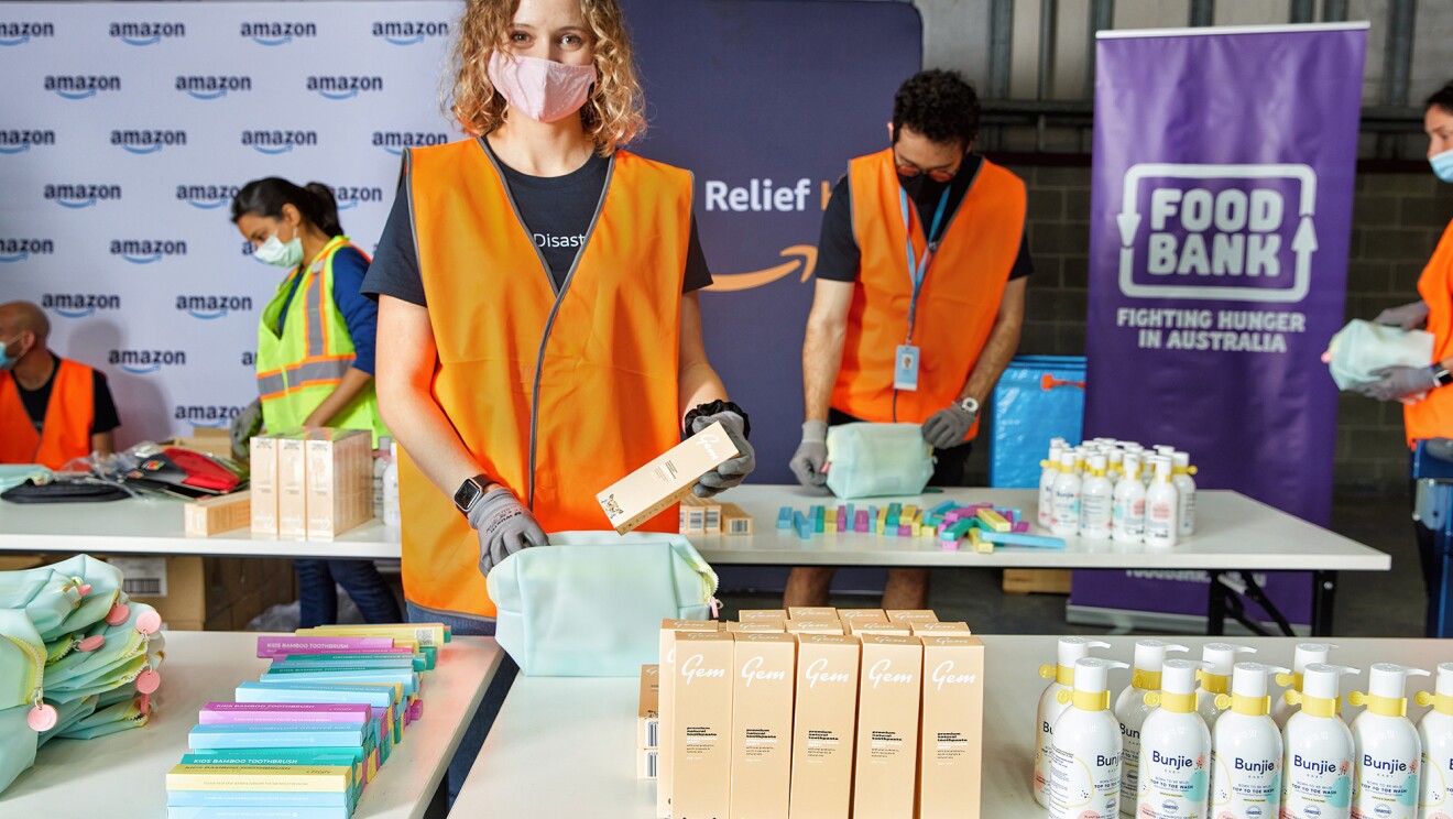 A woman wearing a face mask and orange safety vest packs personal care products into a tote. Behind her, other people wearing safety bests and masks do the same. ope