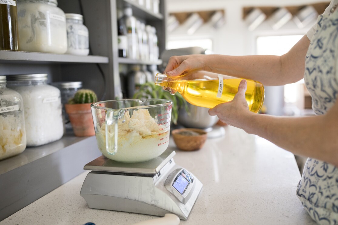 A woman pour's jojoba oil into a large, glass measuring cup on a scale. Other ingredients for soap making are already in measuring cup. Behind the mixture are jars of ingredients for the company's soaps and candles. 