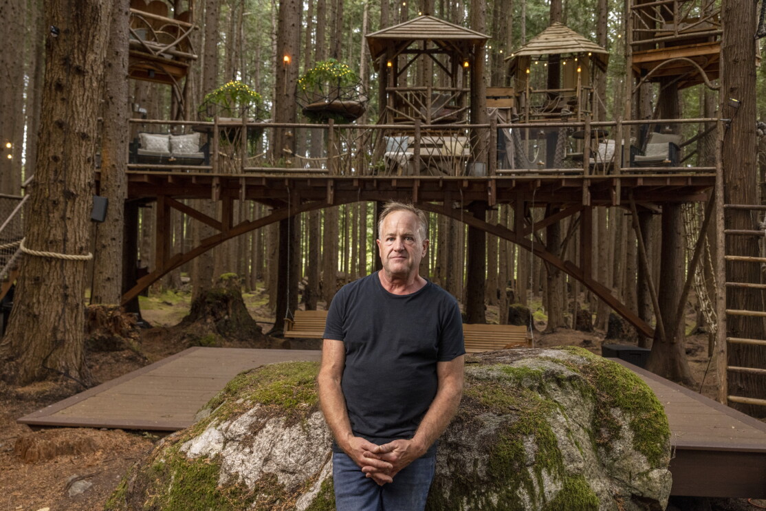 A man stands in front of a large rock, a large wooden structure, and a forest.