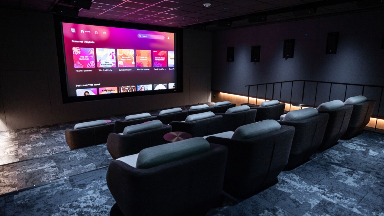 A theater room with a large projected screen at the front and big, soft comfy chairs in three rows. The room is dark with dark blue features and spotlights that can be lit.