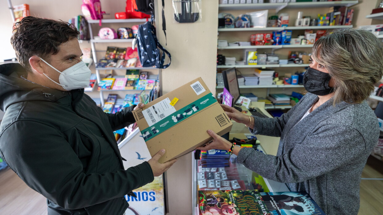 Ana Gallardo regente El Detallito, una papelería y agencia de viajes en Los Corrales, Sevilla. En la papelería Ana entrega un paquete a un cliente. Los dos llevan mascarilla. Ana está detrás del mostrados lleno de libros. En el fondo estanterías llenas de productos de papelería. Ana va con una chaqueta gris, media melena y una mascarilla negra. El cliente va con un anorak negro y una mascarilla blanca y tiene el pelo corto negro. 