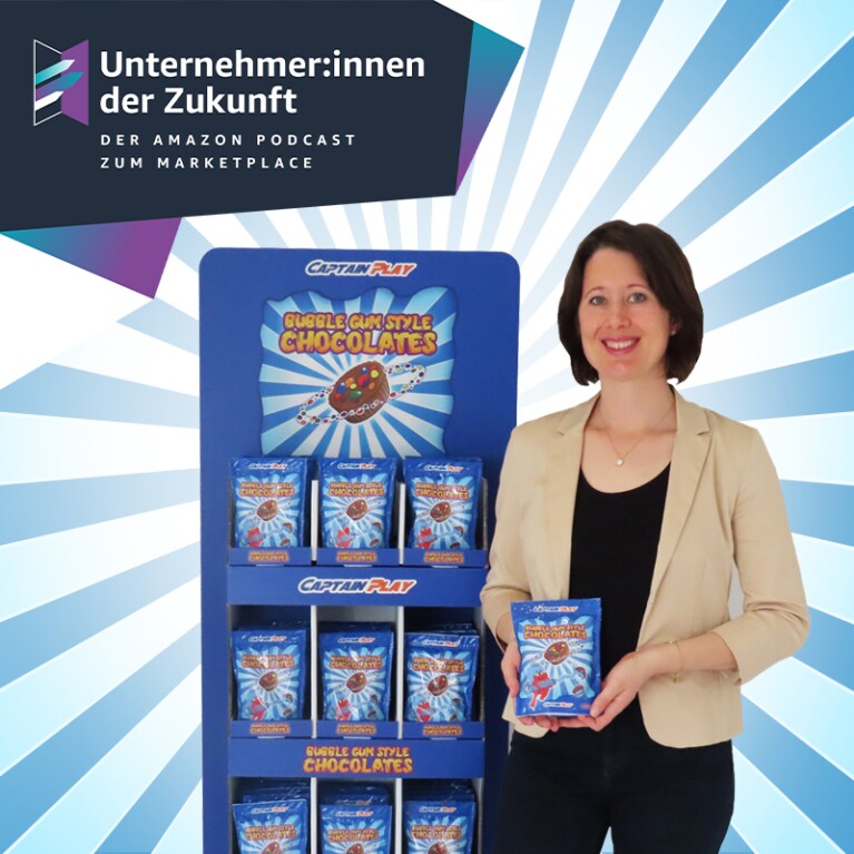 Annika Würtenberger, Gründerin von Captain Play, steht neben einem Süßigkeitenregal. Sie trägt einen beigen Blazer und ein schwarzes Shirt. Sie hat braune, halblange Haare und lächelt in die Kamera. In der Hand hält sie eine Tüte mit Süßigkeiten.