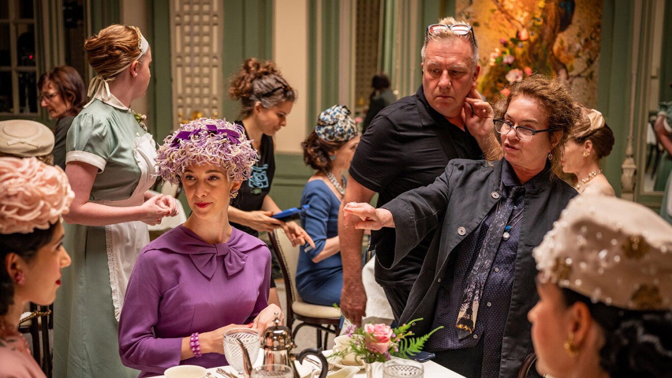 An image of women sitting at a table wearing clothing from the 1950s. A woman points at the costumes giving direction for adjustments.