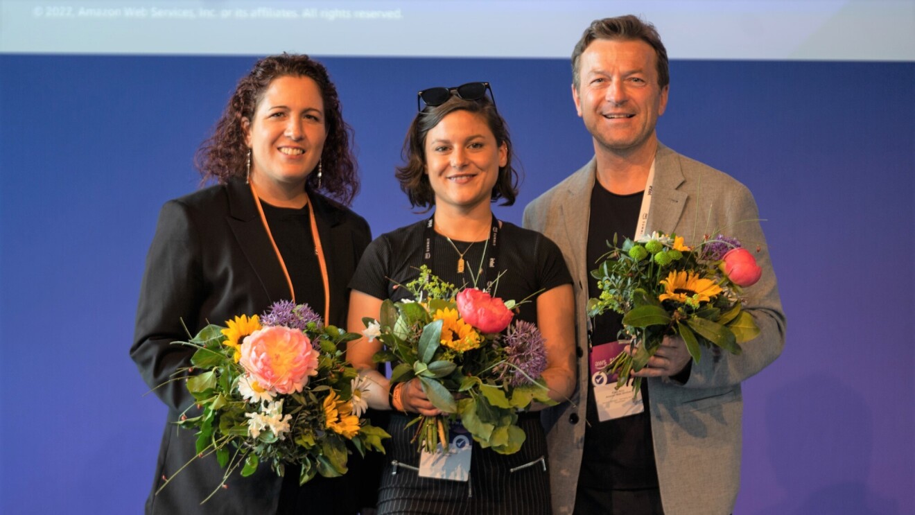 Gruppenbild der AWS-Jury, bestehend aus AWS DACH Chef Klaus Bürg, Softwarearchitektin Nahia Orduna und AWS Community Builder Linda Mohamed, mit Blumensträußen vor einer blauen Wand beim Wettbewerb von AWS GetIT in 2022.