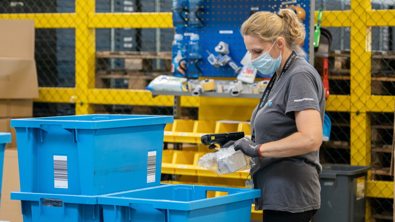 Centro Logístico de Amazon. En una estación logística está una trabajadora de Amazon rubia, con una cola y mascarilla. Camiseta de manga corta de color gris de Prime Day y unas mallas de color negro. Está con unos guantes y un lector de código de barras en la mano derecha. En la mano izquierda está agarrando un producto que está en una bolsa de plástico. Delante de ella tres cajas de color azul. De fondo, una estación con productos que no se pueden apreciar. Y de fondo una malla negra con unas columnas de color amarillo. 