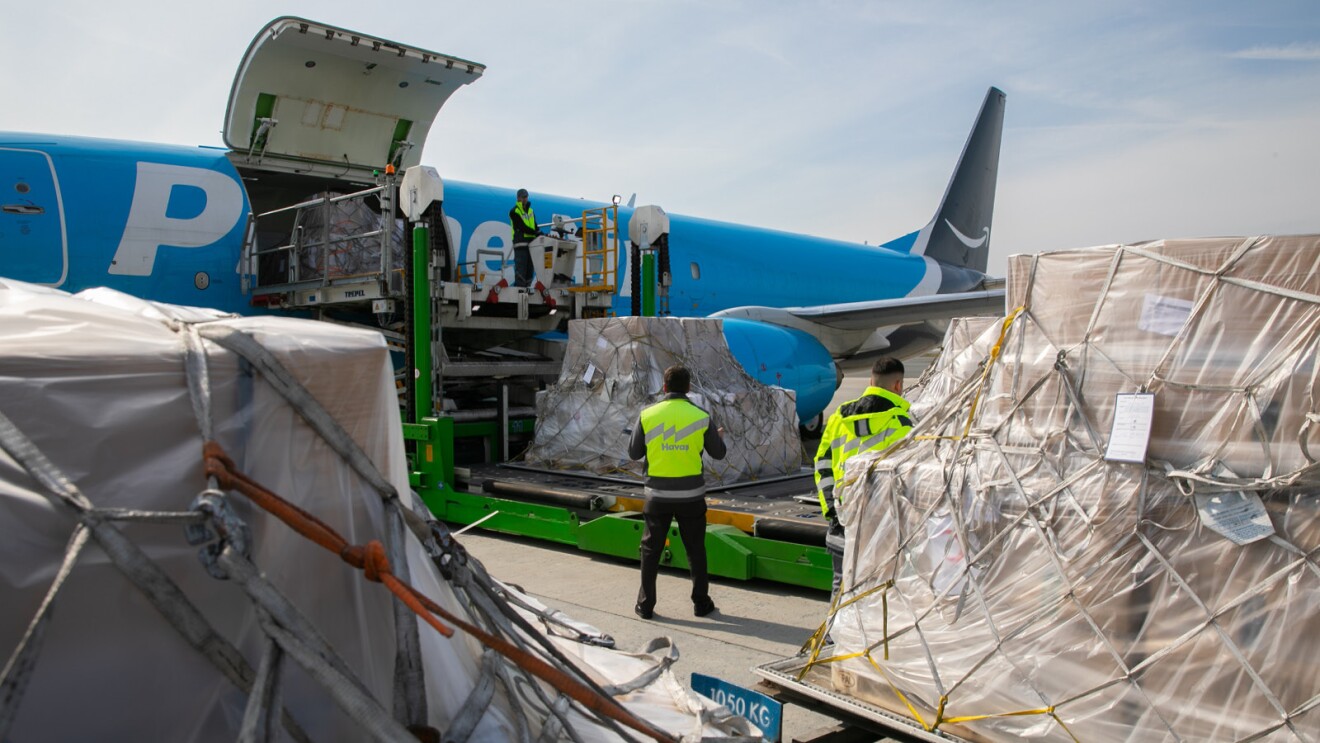 Two men standing among pallets of humanitarian goods, watching as the pallets are loaded on a conveyor belt onto an Amazon Air cargo plane. A third man stand on a lift, ready to accept the pallet onto the plane.