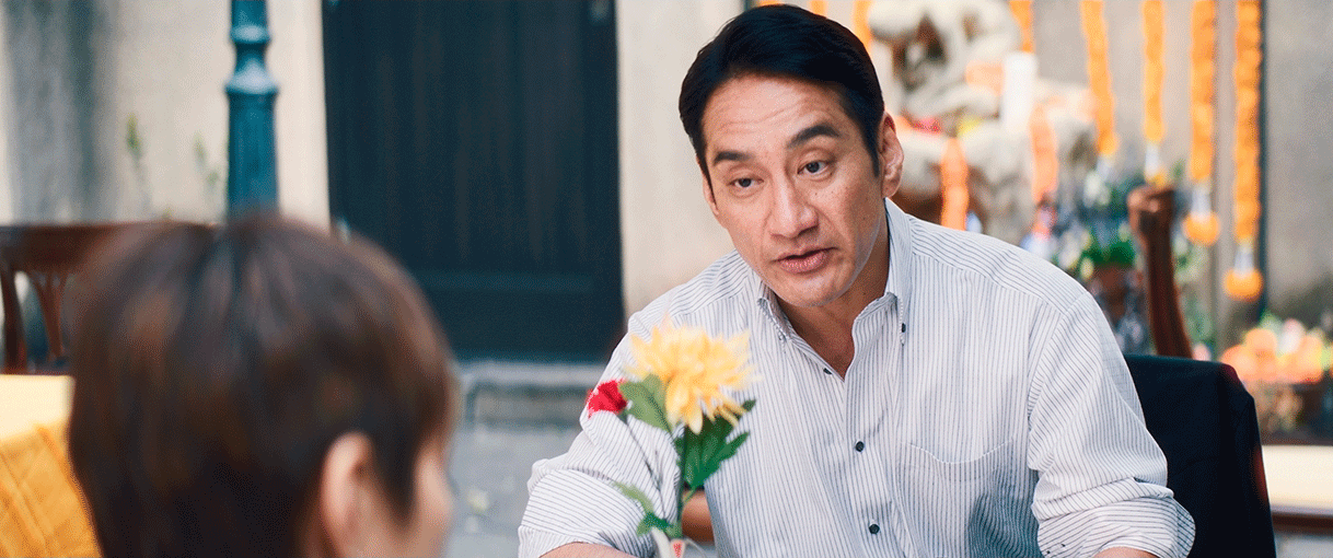 ストライプシャツの中年男性が、手前に写ったショートカットの人物と対峙。男性の前に花がいけられている。後方に壁や扉、街灯、左側にテーブルと椅子が見える。