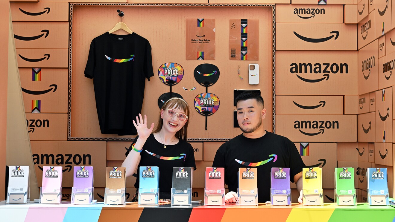 Amazonの段ボールに囲まれたブースの中に黒いTシャツを着た男性と女性がたっている。手前に12色のリーフレットが並び、後ろにはうちわなどが壁に掛けられている