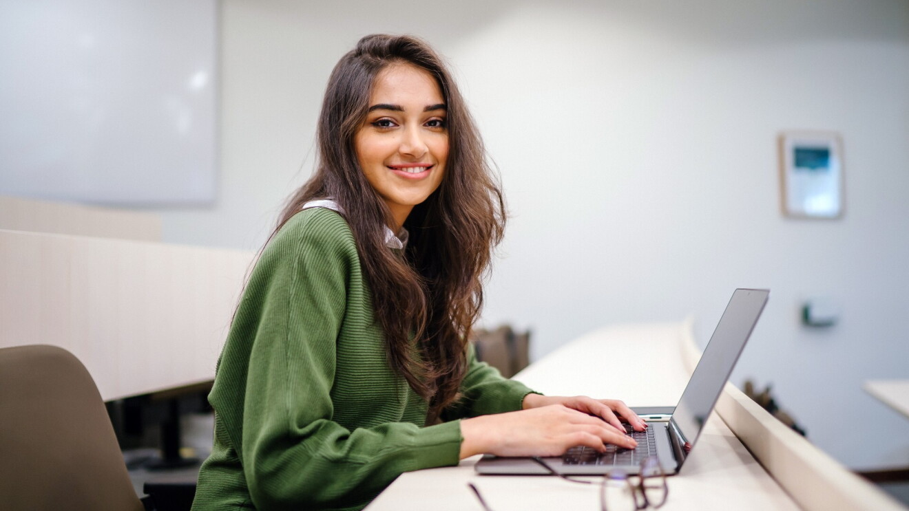 A young woman wearing a green sweatshirt smiles at the camera while studying on her laptop.