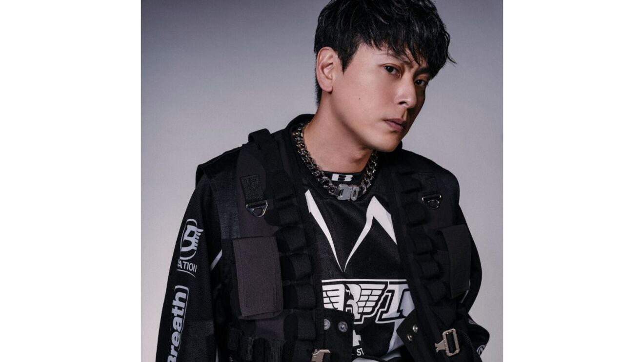 黒い服に黒いアーミーベスト、チェインネックレスを着ている男性（山下健二郎さん）が斜めから険しい表情で正面を見ている