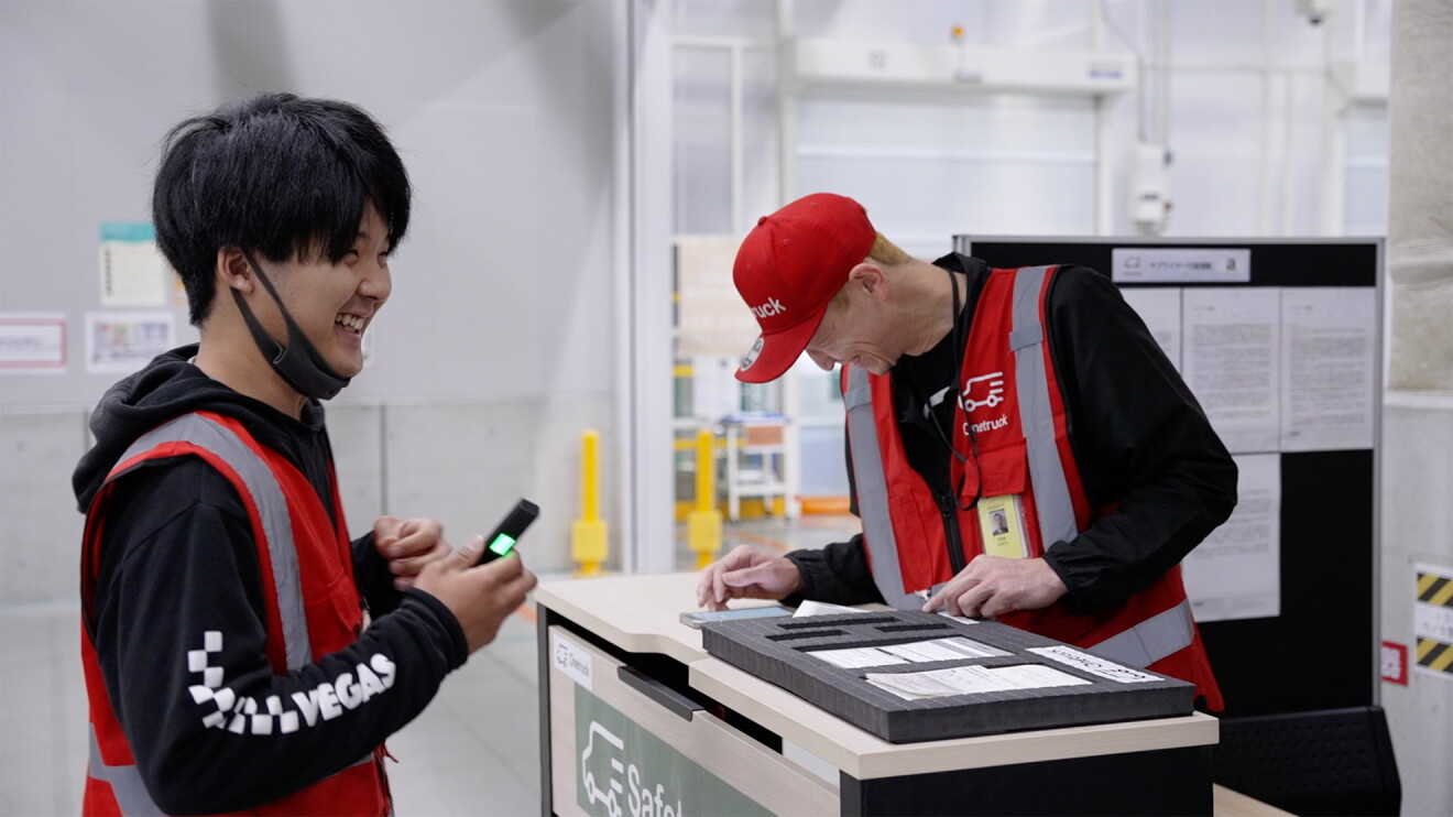 赤い作業用ベストを着た男性2人、一人は作業台の上でスマートフォンを操作している