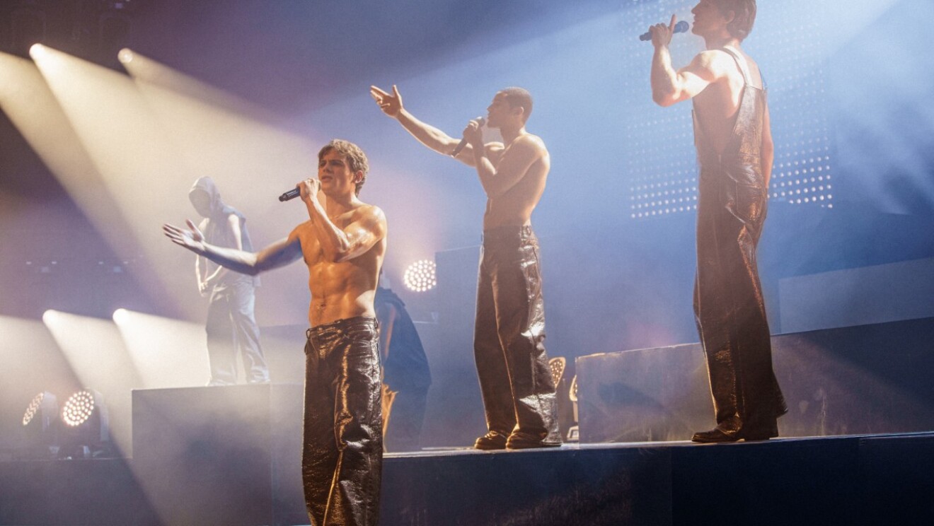 Le boys band se produit torse nu sur une scène illuminée avec un éclairage dramatique.