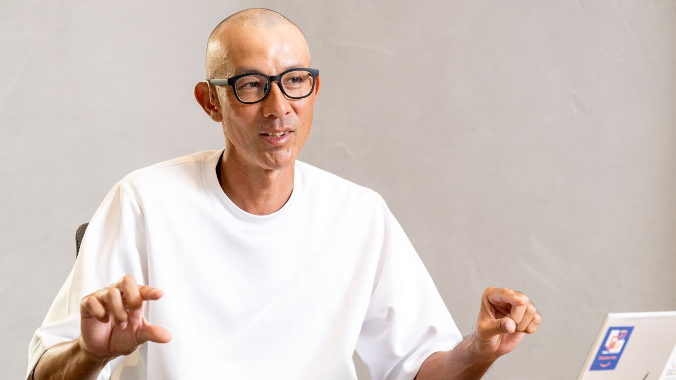 薄いグレーの壁を背景に話をする人物の上半身。黒いフレームの眼鏡をかけ、白いシャツを着ている。両手を軽くあげて人差し指と親指で何か挟むようなジェスチャー。右側にノートパソコンが開いている。