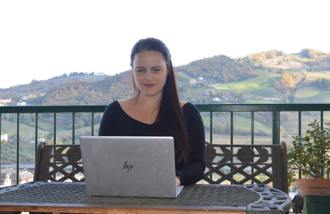 Una donna è seduta ad un tavolo su un terrazzo e ha di fronte a sè un PC. Alle sue spalle delle colline. 