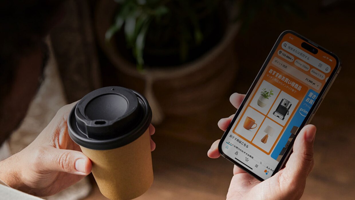 左手に紙製のテイクアウトカップ、右手にスマートホンを持つ人物の手元のショット。スマートホンの画面にはオレンジ色を基調としたAmazonアプリの「おすすめお買い得商品」が表示されている。