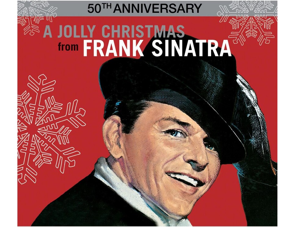 Frank Sinatra wears a suit and white button down, with a rimmed hat on his head, hair slicked, twinkle in his eye. His black gloved hand reaches up to tip his hat. He smiles toward the camera. The background is red, with large illustrated snowflakes falling around him. 