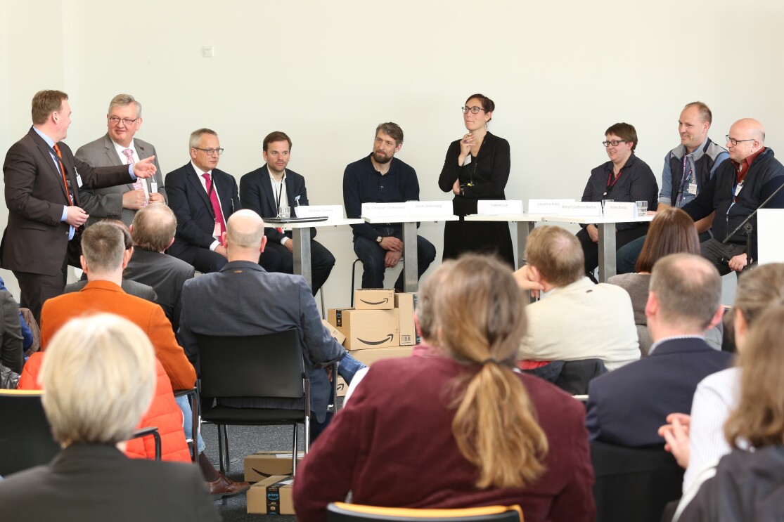 Neun Besucher auf einem Podium, die Gäste sieht man von hinten