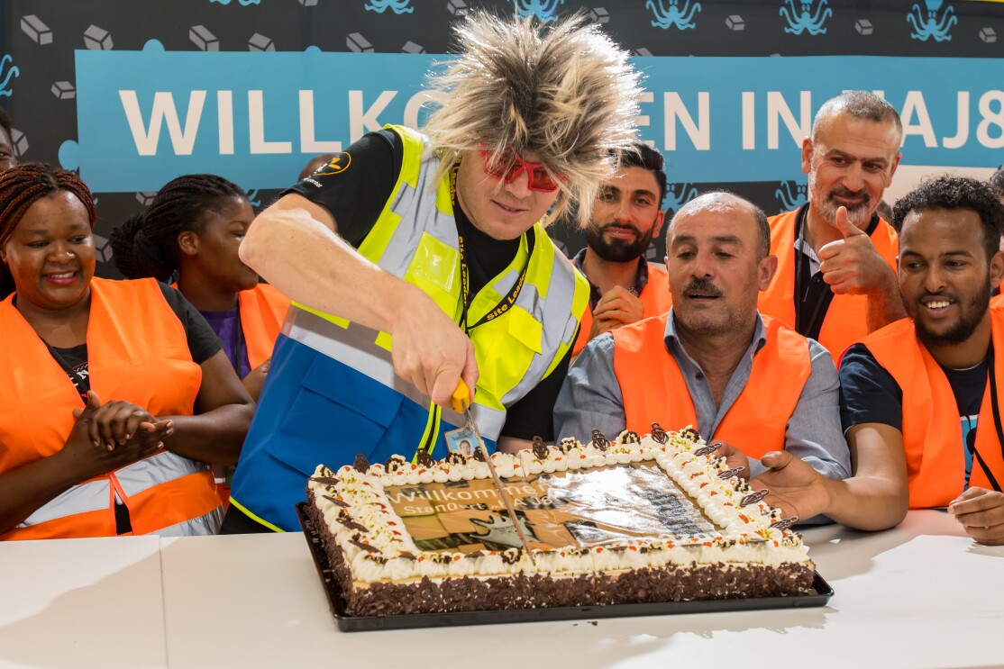 Ein Mann mit Igelperücke und roter Spaßbrille, von Logistikmitarbeitern umgeben, schneidet eine rechteckige Torte an