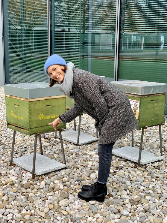 Eine Frau mit blauer Mütze steht an einem grünen Bienenkasten und schaut in die Kamera.